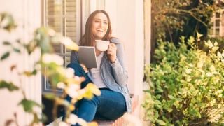 Junge Frau sitzt auf der Fensterbank ihres Eigenheims und trinkt einen Kaffee.